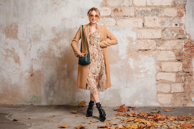 Attraente donna bionda alla moda in cappotto beige che cammina in strada contro il muro d'epoca