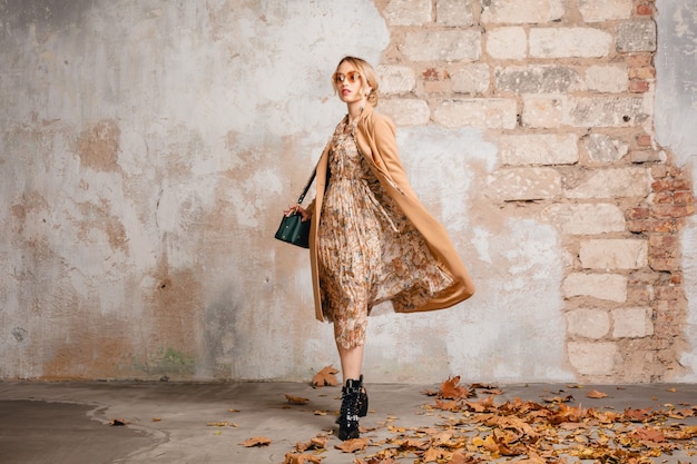 Attraente donna bionda alla moda in cappotto beige che cammina in strada contro il muro d'epoca