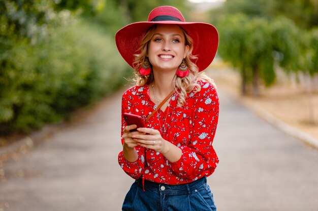 Attraente donna bionda alla moda sorridente in cappello rosso di paglia e camicetta vestito di moda estiva utilizzando il telefono