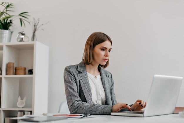 Attraente donna bionda digitando la lettera nel computer portatile al suo posto di lavoro. ritratto di signora in giacca elegante in ufficio luminoso.