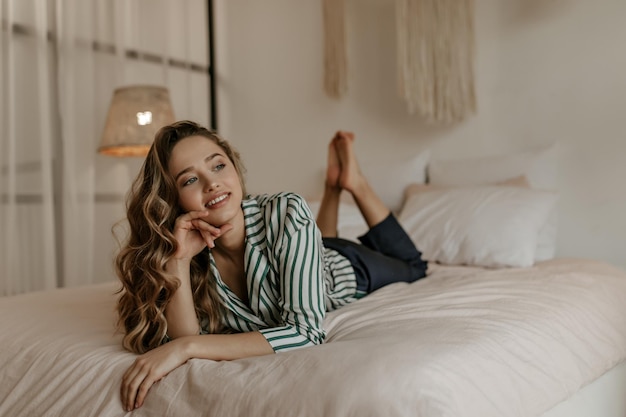 Attraente donna bionda riccia in pantaloni neri e camicia a righe di seta distoglie lo sguardo sorride e riposa