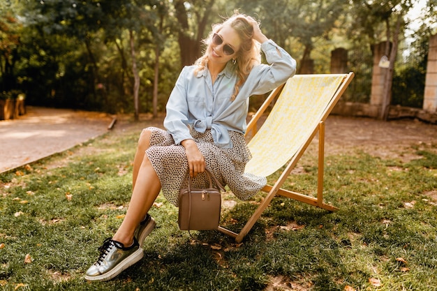 Attraente donna bionda sorridente seduta sulla sedia a sdraio in abito estivo camicia blu e gonna, indossando scarpe da ginnastica argento, occhiali da sole eleganti e borsa, stile di moda di strada