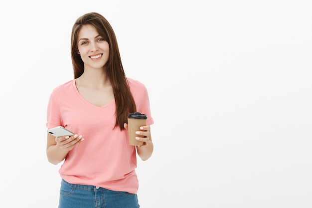 Attraente donna bruna sorridente in posa in studio con il suo telefono e caffè