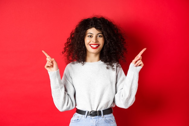 Attraente donna caucasica che mostra modo, puntando le dita lateralmente su due promo, dimostrando varianti e sorridendo, in piedi su sfondo rosso.