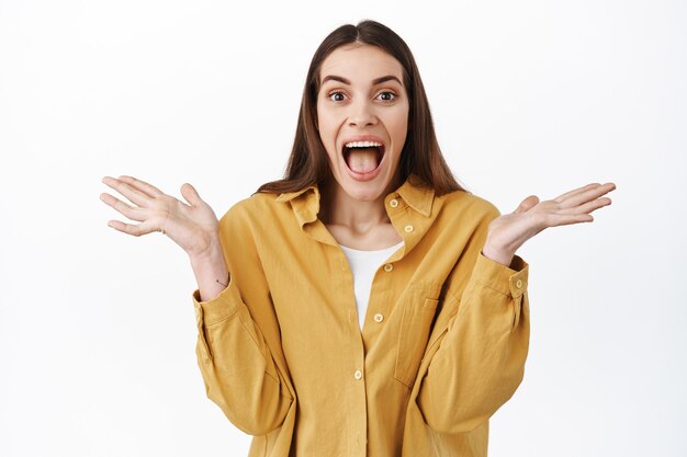 Attraente donna caucasica urla di stupore e sorpresa, allarga le mani di lato e grida, reazione su un nuovo super cool, gioia e celebrazione, in piedi sul muro bianco