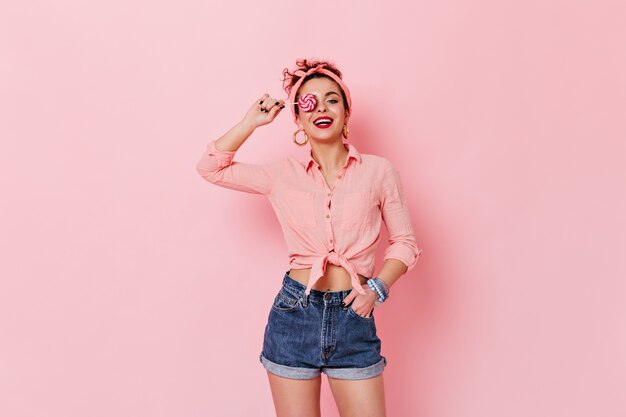 Attraente donna che indossa la camicia pin-up e la fascia in posa con lecca-lecca sullo spazio rosa.