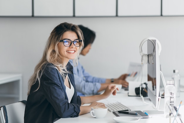 Attraente donna che ride libero professionista in posa con una tazza di caffè al suo posto di lavoro. Studente cinese in camicia blu lavora con il documento nel campus con un'amica bionda con gli occhiali.