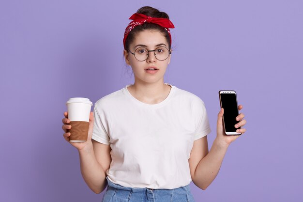 Attraente donna con espressione facciale stupita, tiene il cellulare con schermo vuoto e caffè da portare via
