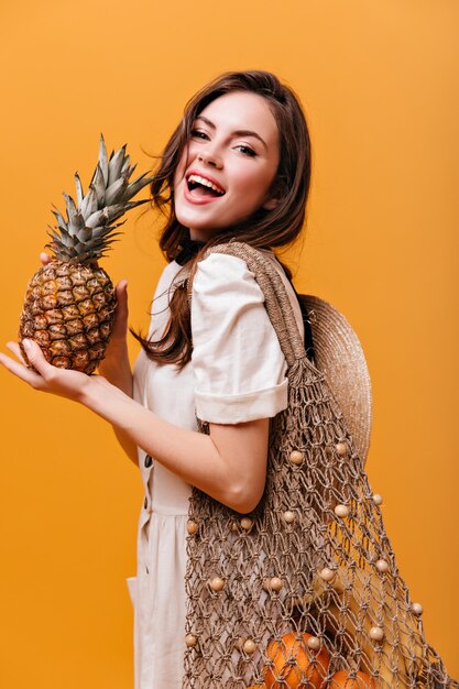 Attraente donna dai capelli lunghi in abito bianco in posa con la borsa della spesa lavorata a maglia e tenendo l'ananas.