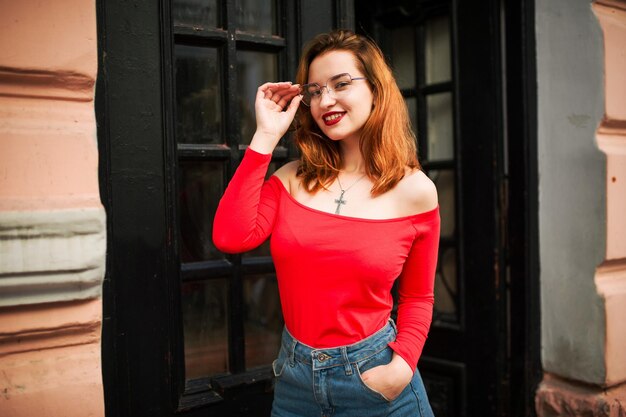 Attraente donna dai capelli rossi in occhiali da vista indossare su camicetta rossa e gonna di jeans in posa in strada contro la vecchia porta di legno