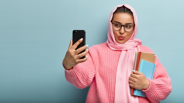 Attraente donna di razza mista con i capelli scuri, indossa copricapo di seta, tiene il cellulare davanti, scatta un selfie, pubblica un post nei social network, tiene due taccuini a spirale, prende il videomessaggio