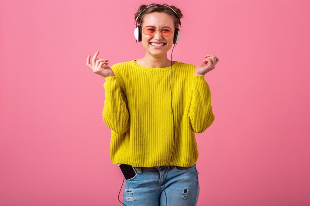 Attraente donna divertente felice che ascolta la musica in cuffie vestita in abito stile colorato hipster isolato sulla parete rosa, indossa un maglione giallo e occhiali da sole, divertendosi