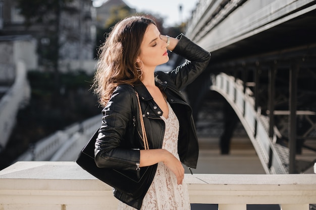 Attraente donna elegante che cammina in strada in abito alla moda, con borsa in pelle scamosciata, indossa giacca di pelle nera e abito di pizzo bianco, stile primavera autunno