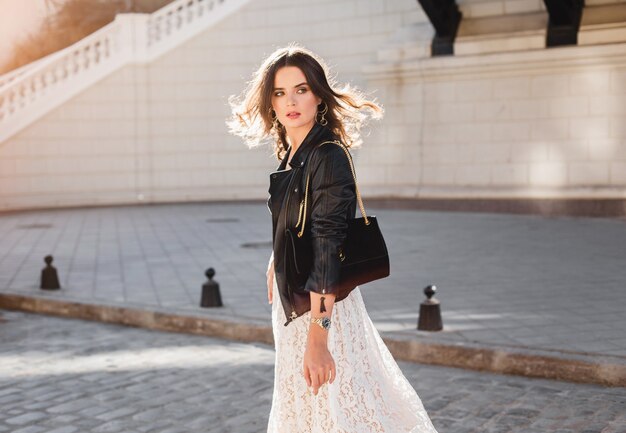 Attraente donna elegante che cammina per strada in abito alla moda, tenendo la borsa in pelle scamosciata, indossa una giacca di pelle nera e abito di pizzo bianco, stile primavera autunno, girandosi in movimento