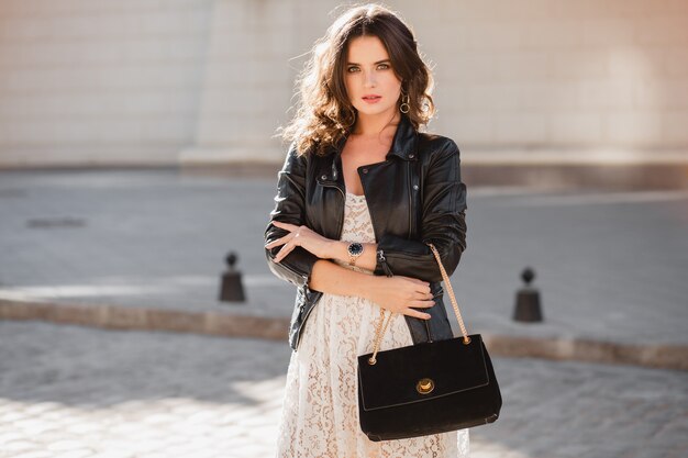 Attraente donna elegante che cammina per strada in abito alla moda, tenendo la borsa, indossa una giacca di pelle nera e abito di pizzo bianco, stile primavera autunno