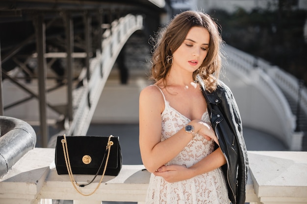 Attraente donna elegante che cammina per strada in abito alla moda, tenendo la borsa, indossa una giacca di pelle nera e abito di pizzo bianco, stile primavera autunno