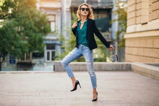 Attraente donna elegante che cammina per una strada cittadina con scarpe col tacco alto, jeans blu wesaring, giacca nera, camicetta verde, occhiali da sole, borsetta con in mano, tendenza della moda dell'estate, bella signora magra