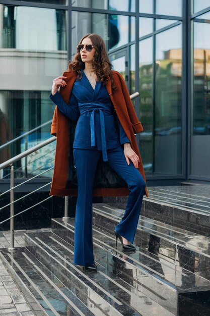 Attraente donna elegante con passeggiate in strada di affari della città urbana vestita di caldo cappotto marrone e abito blu, stile di strada moda alla moda primavera autunno, indossando occhiali da sole