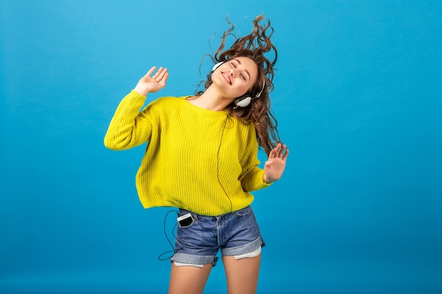 Attraente donna felice sorridente ballando ascoltando musica in cuffia in abito elegante hipster isolato su sfondo blu studio, indossando pantaloncini e maglione giallo