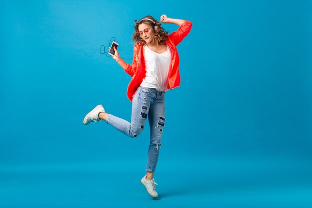 Attraente donna felice sorridente che balla ascoltando musica in cuffie vestita in abito stile hipster isolato su sfondo blu studio, indossando occhiali da sole e giacca rosa