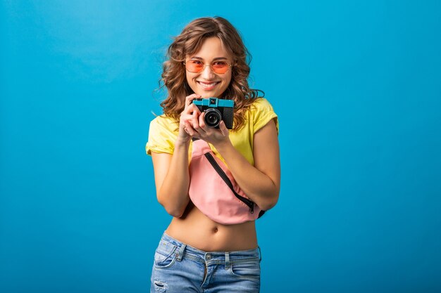 Attraente donna felice sorridente in posa con la macchina fotografica d'epoca scattare foto vestita in abito colorato estate hipster isolato su sfondo blu studio