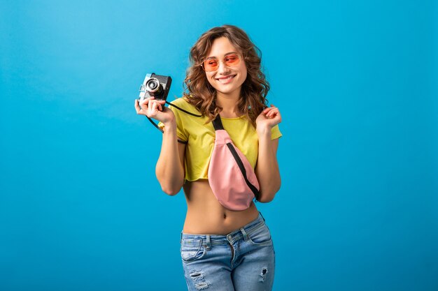 Attraente donna felice sorridente in posa con la macchina fotografica d'epoca scattare foto vestita in abito colorato estate hipster isolato su sfondo blu studio