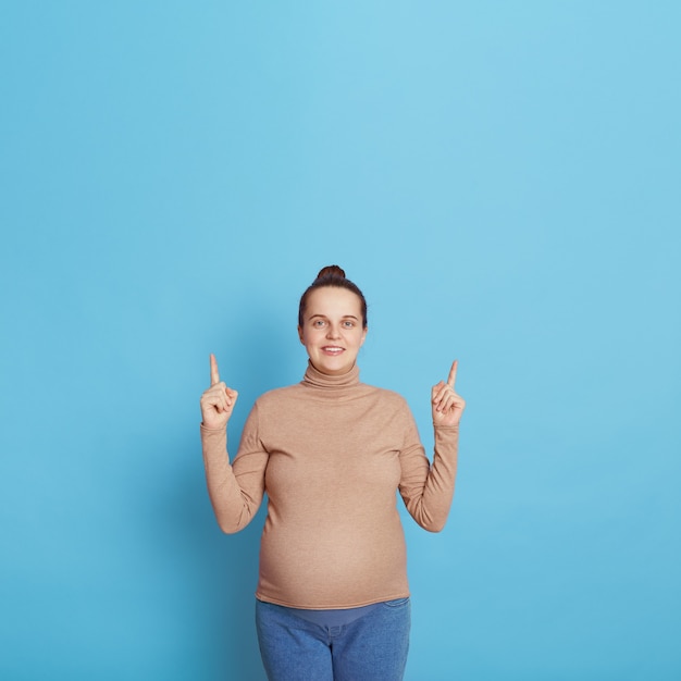 Attraente donna incinta europea che punta il dito verso l'alto con entrambi gli indici che indossano abbigliamento casual, indicando qualcosa sopra la sua testa, copia spazio per le promozioni.