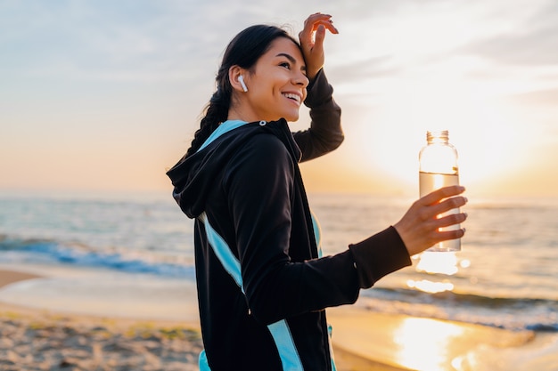 Attraente donna magra che fa esercizi sportivi sulla spiaggia all'alba del mattino in abbigliamento sportivo, acqua potabile assetata in bottiglia, stile di vita sano, ascolto di musica con auricolari wireless, sensazione di caldo