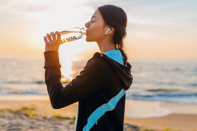 Attraente donna magra facendo esercizi sportivi sulla spiaggia all'alba del mattino in abbigliamento sportivo, acqua potabile assetata in bottiglia, stile di vita sano, ascolto di musica su auricolari wireless, sorridendo felice