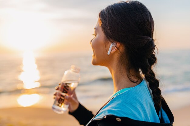 Attraente donna magra facendo esercizi sportivi sulla spiaggia all'alba del mattino in abbigliamento sportivo, acqua potabile assetata in bottiglia, stile di vita sano, ascolto di musica su auricolari wireless, sorridendo felice