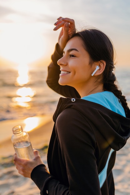 Attraente donna magra facendo esercizi sportivi sulla spiaggia di alba mattutina in abbigliamento sportivo, acqua potabile assetata in bottiglia, stile di vita sano, ascolto di musica con auricolari wireless, calda giornata estiva