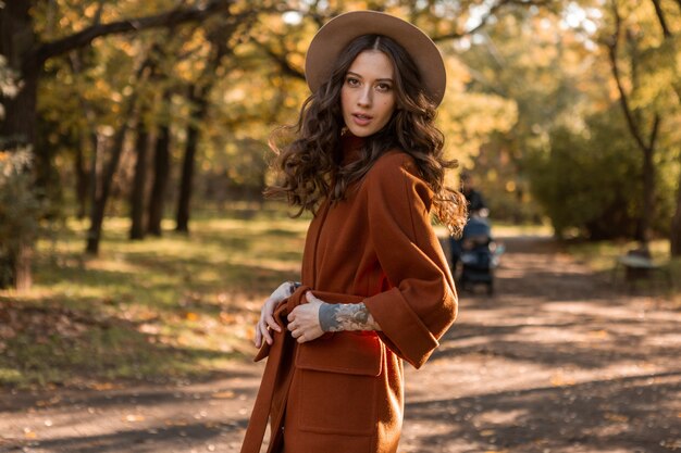 Attraente donna magra sorridente elegante con capelli ricci che cammina nel parco vestito con un cappotto marrone caldo, stile di strada moda alla moda autunno