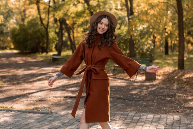 Attraente donna magra sorridente elegante con capelli ricci che cammina nel parco vestito con un cappotto marrone caldo, stile di strada moda alla moda autunno