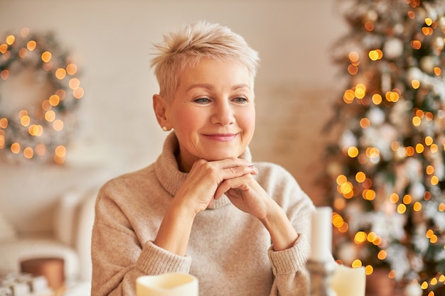 Attraente donna matura in atmosfera festosa anticipando il nuovo anno pensando ai regali per la famiglia, seduto in soggiorno, circondato da albero di Natale decorato, ghirlande e luci di ghirlanda