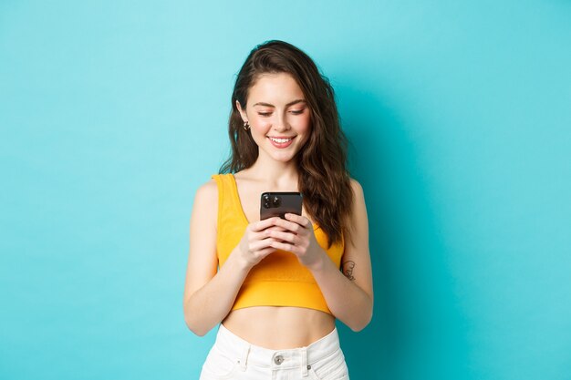 Attraente donna moderna in chat sul telefono cellulare, guardando lo schermo con un sorriso tenero, messaggistica su app di appuntamenti, in piedi su sfondo blu.