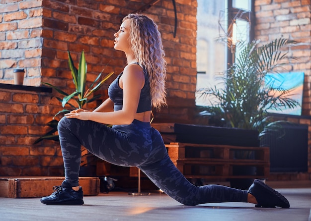 Attraente donna positiva per il corpo sta facendo esercizio aerobico al loft moderno. Indossa leggings sportivi, reggiseno e scarpe da ginnastica. La donna ha i capelli ricci biondi.