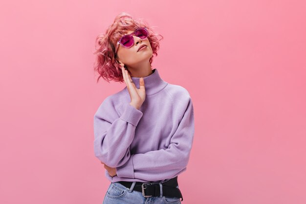 Attraente donna riccia in maglione di cashmere viola e occhiali da sole fucsia posa sul muro isolato