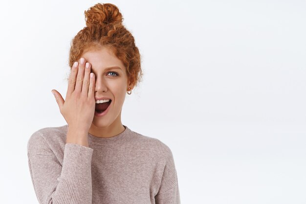 Attraente donna rossa sfacciata con capelli ricci pettinati, copre un lato del viso, mostrando la mano, nuova manicure, sorridente stupito, muro bianco