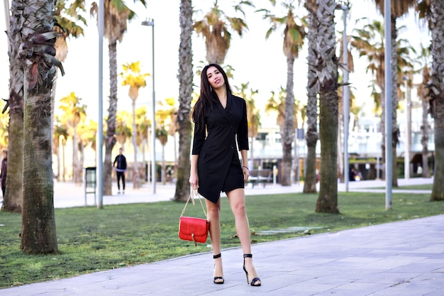 Attraente donna sicura di sé sexy che cammina per strada con palme, vestito nero minimalista da donna di affari moderna, viaggiando in Spagna.