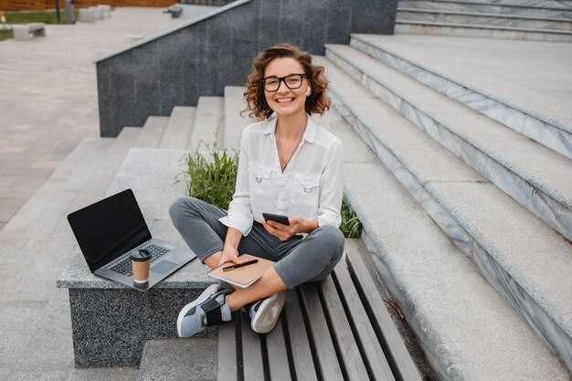 Attraente donna sorridente alla moda con gli occhiali che lavora digitando sul laptop