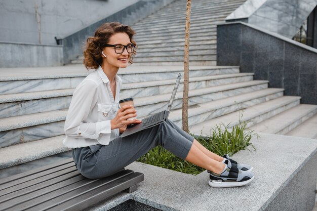 Attraente donna sorridente alla moda con gli occhiali che lavora digitando sul laptop