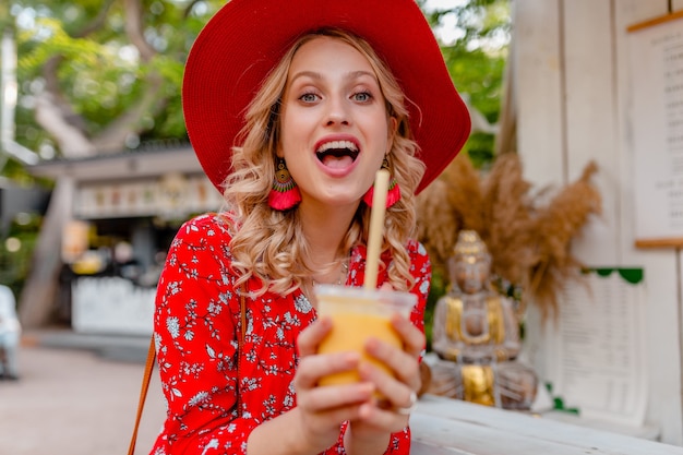 Attraente donna sorridente bionda elegante in cappello rosso di paglia e camicetta vestito di moda estiva bere frullato cocktail di frutta naturale