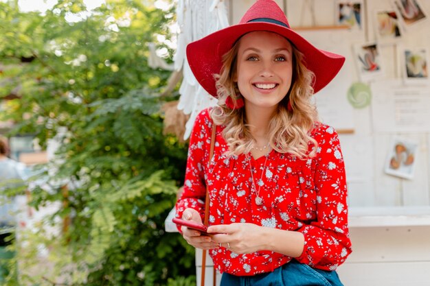 Attraente donna sorridente bionda elegante in cappello rosso di paglia e camicetta vestito di moda estiva che tiene utilizzando smart phone cafe