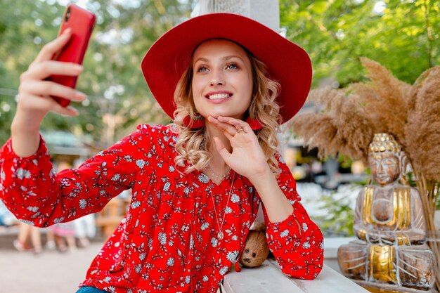 Attraente donna sorridente bionda elegante in cappello rosso di paglia e camicetta vestito di moda estiva prendendo foto selfie sul caffè smartphone fotocamera