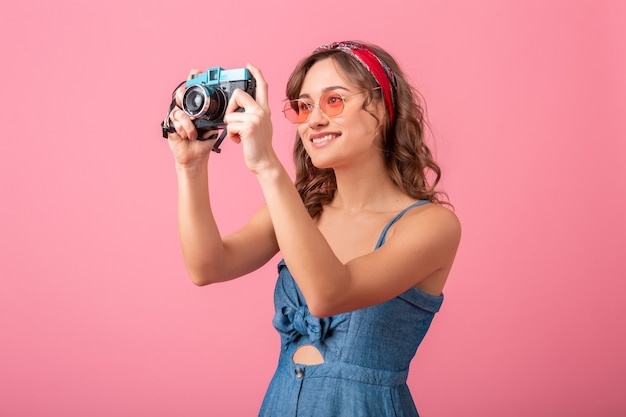 Attraente donna sorridente che cattura foto sulla fotocamera vintage indossando abiti in denim e occhiali da sole, isolati su sfondo rosa