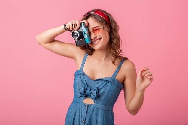 Attraente donna sorridente che cattura foto sulla fotocamera vintage, puntando il dito verso l'alto, indossando abiti di jeans e occhiali da sole isolati su sfondo rosa