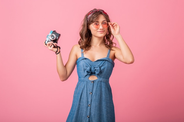 Attraente donna sorridente che cattura foto sulla fotocamera vintage, puntando il dito verso l'alto, indossando abiti di jeans e occhiali da sole isolati su sfondo rosa