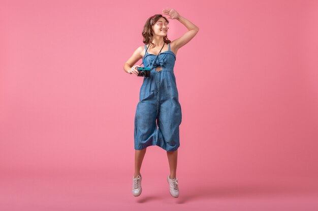 Attraente donna sorridente che salta attivo prendendo foto sulla fotocamera vintage che indossa un abito denim isolato su sfondo rosa