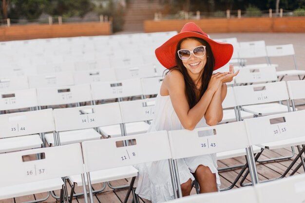 Attraente donna sorridente felice vestita in abito bianco, cappello rosso, occhiali da sole seduti in un teatro all'aperto estivo da solo, molte sedie, tendenza della moda di strada primaverile, ciao agitando la mano