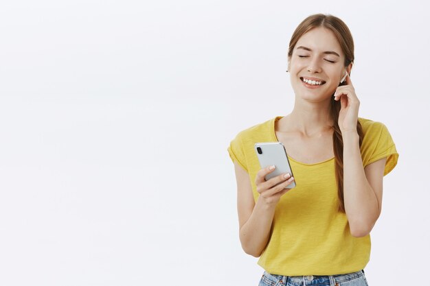 Attraente donna sorridente in cuffia ascoltando musica o podcast, utilizzando lo smartphone